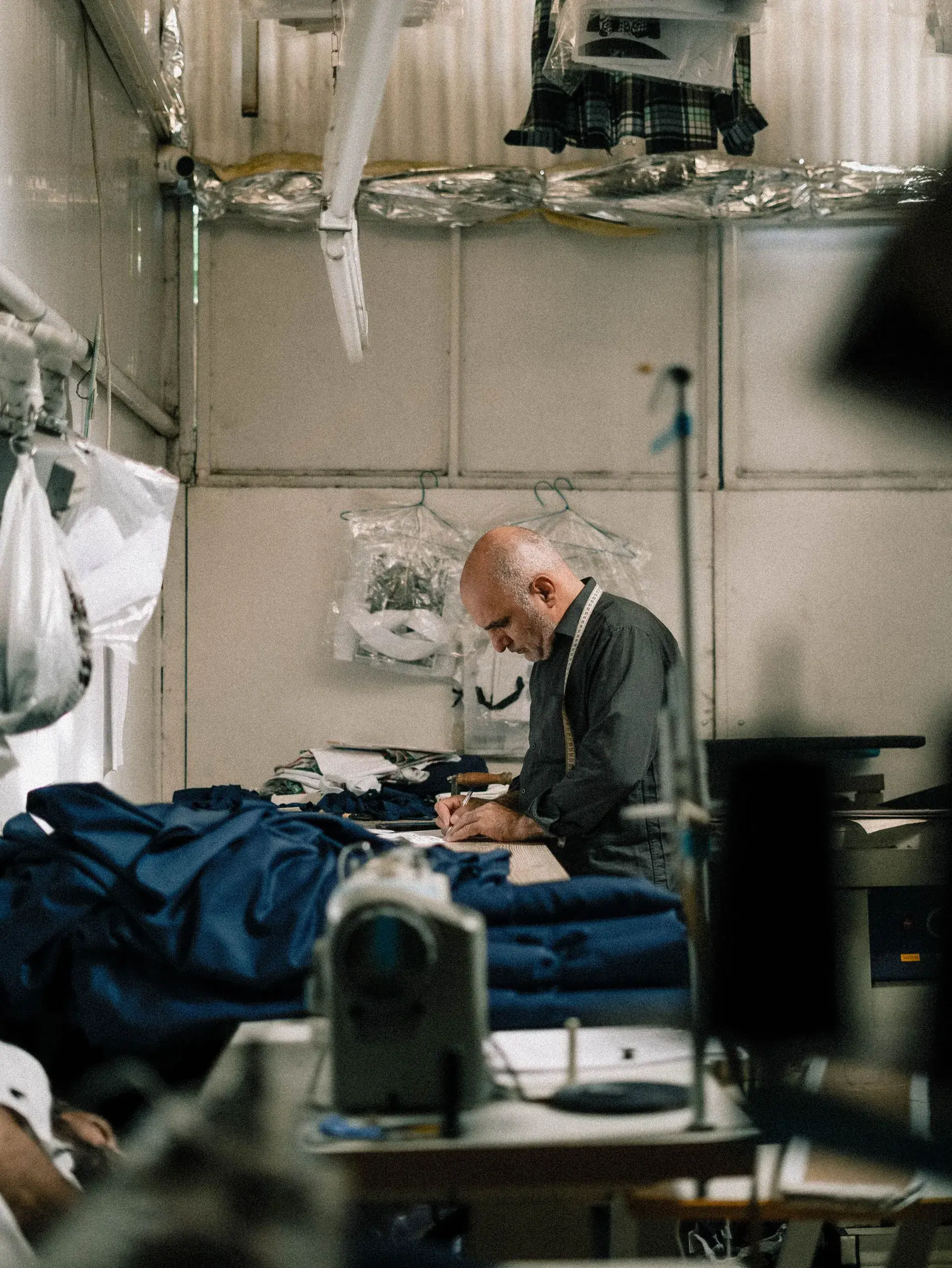 Tailor with a tape measure writing at a workbench beside folded navy garments, with a sewing machine in the foreground — stock illustration, factory photo coming soon